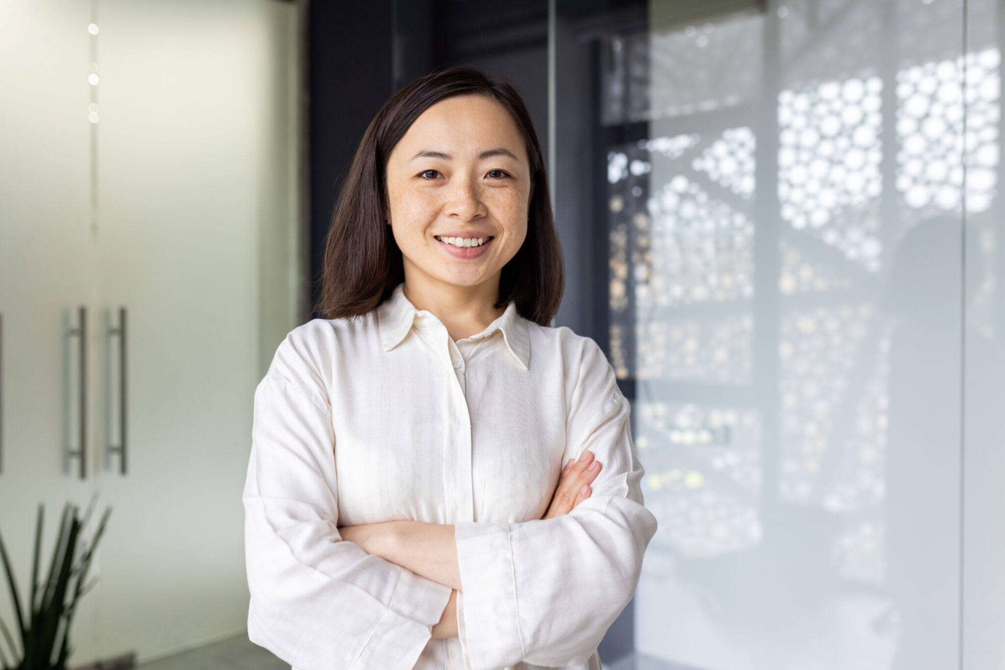 Portrait of young mature asian businesswoman entrepreneur, woman with crossed arms smiling and looking at camera, working inside office at workplace, standing near window in light clothes